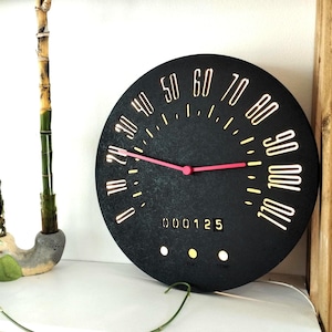 Könnte beinhalten: Schwarze Uhr mit einem Tachodesign. Das Zifferblatt der Uhr hat weiße Zahlen und einen roten Zeiger. Das Zifferblatt der Uhr hat auch eine digitale Anzeige, die "000125" anzeigt.