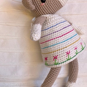 May include: A crocheted stuffed animal fox wearing a white dress with colorful stripes and embroidered flowers. The fox has a brown and white body with black eyes and a black nose.