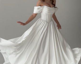 May include: A white off-the-shoulder wedding dress with a fitted bodice and a flowing skirt. The dress has a sweetheart neckline and short puffed sleeves.