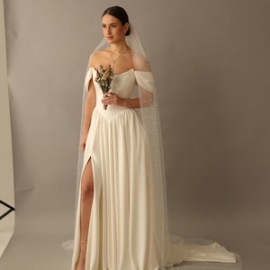 May include: An off-white floor-length gown featuring an off-the-shoulder neckline, a fitted bodice, and a skirt with a high side slit. A long, sheer veil embellished with scattered white pearls is worn over the hair, extending to the floor.