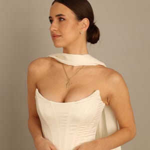 May include: A cream-colored strapless dress with a fitted bodice and flowing skirt. A gold cross pendant hangs from a delicate chain. The model is also wearing a pair of small, round earrings. The dress has a cape-like feature.