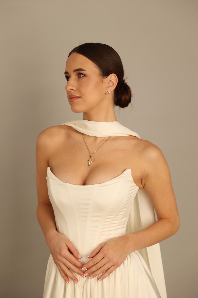 May include: A cream-colored strapless dress with a fitted bodice and flowing skirt. A gold cross pendant hangs from a delicate chain. The model is also wearing a pair of small, round earrings. The dress has a cape-like feature.