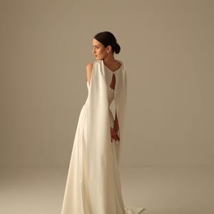 Pu&ograve; includere: Un abito da sposa bianco con una gonna fluida e un mantello sovrapposto. L'abito presenta una schiena a goccia e un corpetto aderente. La modella &egrave; girata di lato, mostrando il design e la silhouette dell'abito. Il tessuto appare liscio ed elegante.