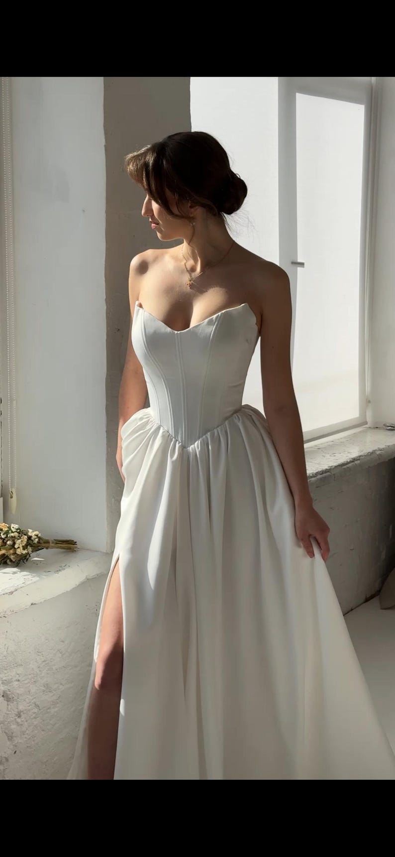 May include: A white strapless wedding dress with a fitted bodice and a flowing skirt. The skirt has a high slit on the left side and a gathered detail at the waist.