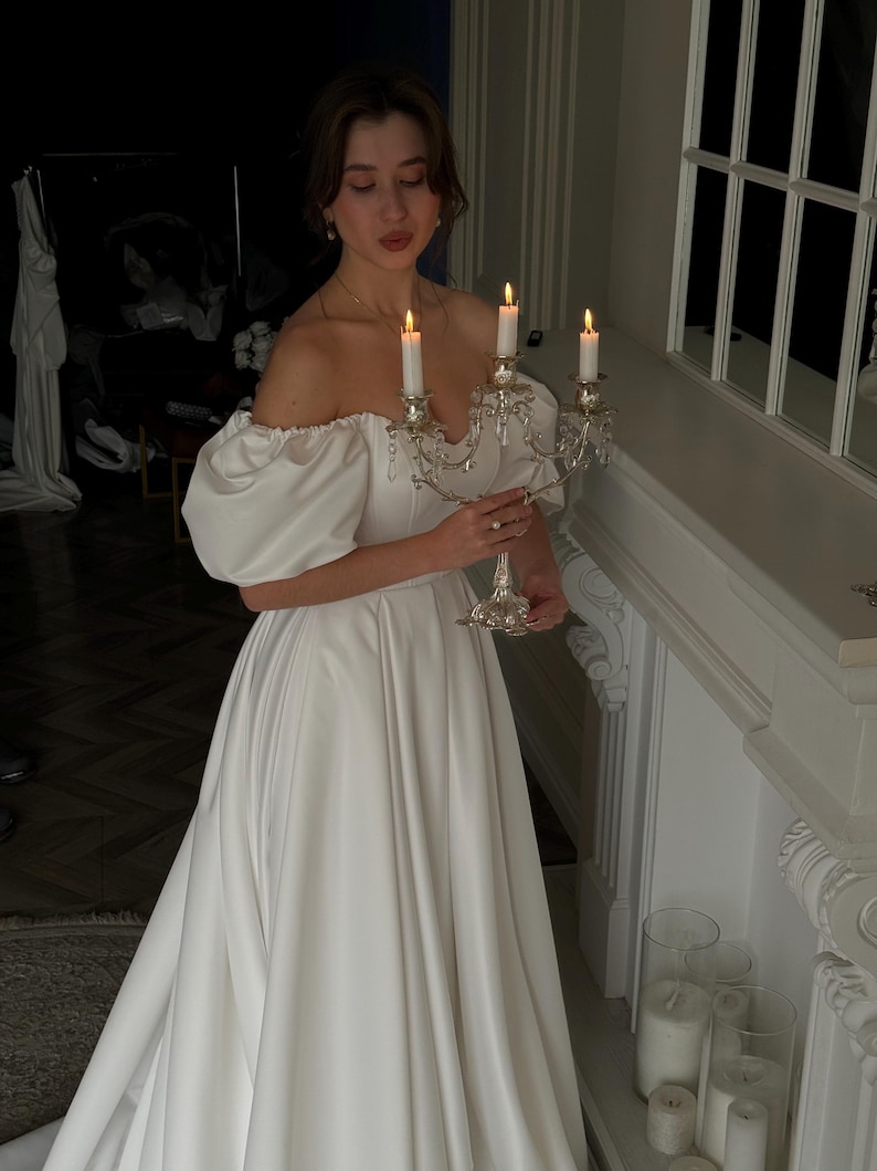 May include: A white off-the-shoulder satin a-line wedding dress with puff sleeves. The dress has a fitted bodice and a flowing skirt.  A silver candelabra with lit candles is held in front of the dress.