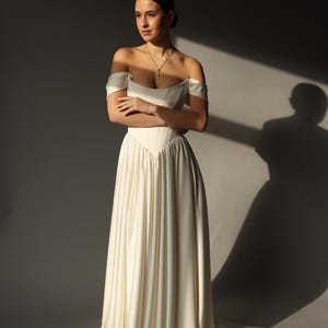 May include: An off-white, off-the-shoulder wedding dress with a fitted bodice and flowing skirt. The dress features a sweetheart neckline and a subtle train. A delicate necklace with a cross pendant is visible.