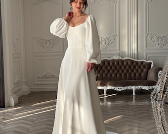 May include: A white wedding dress with a fitted bodice and a flowing skirt. The dress has long sleeves with puffed shoulders and a square neckline.