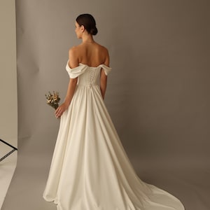 May include: A white off-the-shoulder wedding dress with a long train. The dress features a fitted bodice with a lace-up back and a flowing skirt. The bride is holding a small bouquet of dried flowers.