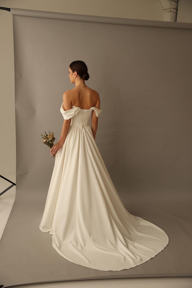 May include: A white off-the-shoulder wedding dress with a long train. The dress features a fitted bodice with a lace-up back and a flowing skirt. The bride is holding a small bouquet of dried flowers.