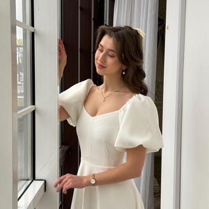 May include: A white dress with a square neckline and puffed sleeves. The dress has a fitted bodice and a flowing skirt.