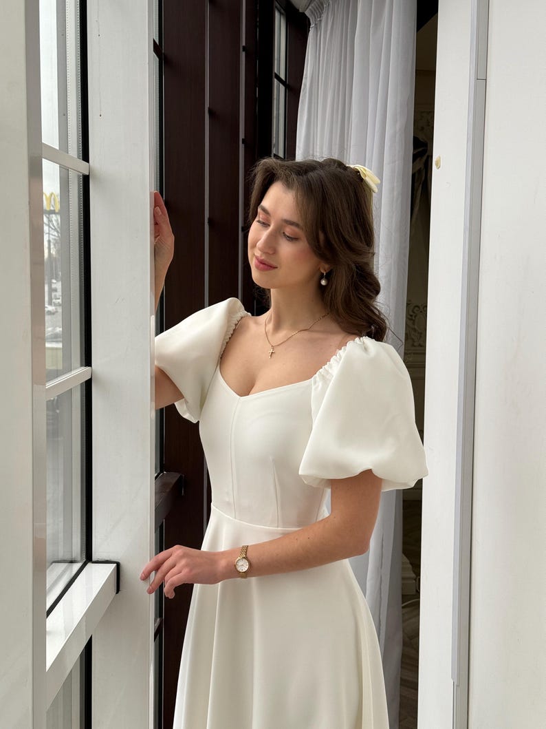 May include: A white dress with a square neckline and puffed sleeves. The dress has a fitted bodice and a flowing skirt.