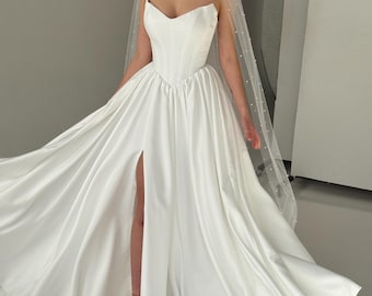 May include: A white strapless wedding dress with a corset-style bodice, a full floor-length skirt featuring a high front slit, and a long sheer veil adorned with small white pearls. The gown is made of a smooth, satin-like fabric.