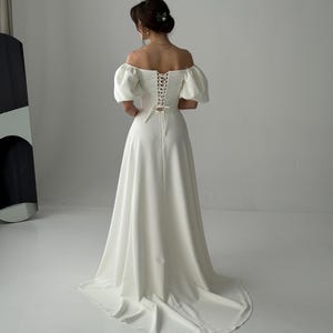 May include: A white off-the-shoulder wedding dress with a fitted bodice and a full skirt. The bodice has a lace-up back and the sleeves are puffed. The dress has a train.