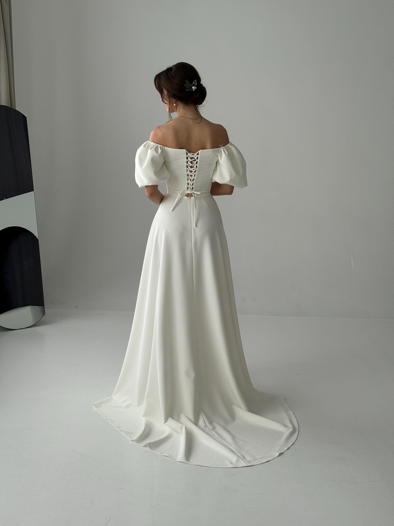 May include: A white off-the-shoulder wedding dress with a fitted bodice and a full skirt. The bodice has a lace-up back and the sleeves are puffed. The dress has a train.