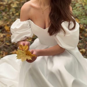 May include: White off-the-shoulder satin a-line wedding dress with puffed sleeves. The dress has a sweetheart neckline and a floor-length skirt. The fabric is smooth and has a subtle sheen. The dress is shown on a model sitting on the ground in a forest setting.