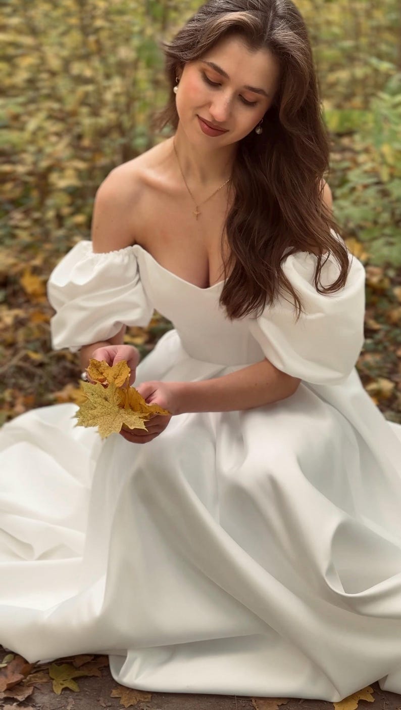May include: White off-the-shoulder satin a-line wedding dress with puffed sleeves. The dress has a sweetheart neckline and a floor-length skirt. The fabric is smooth and has a subtle sheen. The dress is shown on a model sitting on the ground in a forest setting.