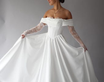 May include: A white off-the-shoulder wedding dress with long lace sleeves. The dress features a fitted bodice and a flowing skirt. A gold cross necklace is visible. The model is wearing a bun.
