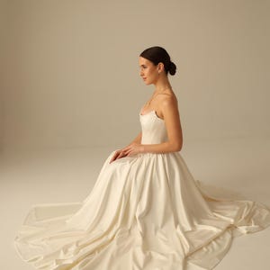 May include: A strapless ivory wedding gown with a fitted bodice and a flowing skirt. The dress features a sweetheart neckline and a long train. The model is seated, showcasing the dress's elegant design and fabric.