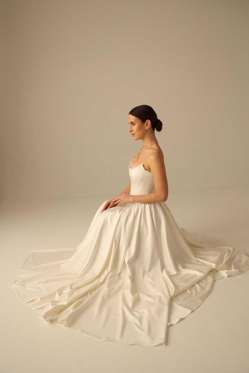 May include: A strapless ivory wedding gown with a fitted bodice and a flowing skirt. The dress features a sweetheart neckline and a long train. The model is seated, showcasing the dress's elegant design and fabric.