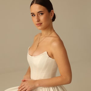 May include: A strapless ivory satin wedding gown with a fitted bodice and full skirt. The model is wearing a gold necklace. The gown is elegant and classic, perfect for a special occasion.