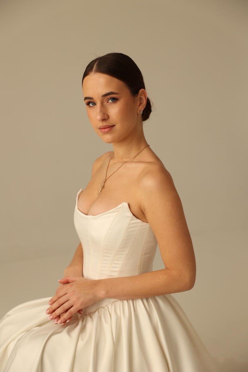 May include: A strapless ivory satin wedding gown with a fitted bodice and full skirt. The model is wearing a gold necklace. The gown is elegant and classic, perfect for a special occasion.