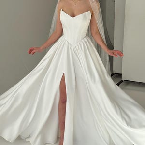 May include: A white strapless wedding dress with a sweetheart neckline and a thigh-high slit. The dress has a fitted bodice and a flowing skirt. The dress is paired with a white veil with pearl embellishments.