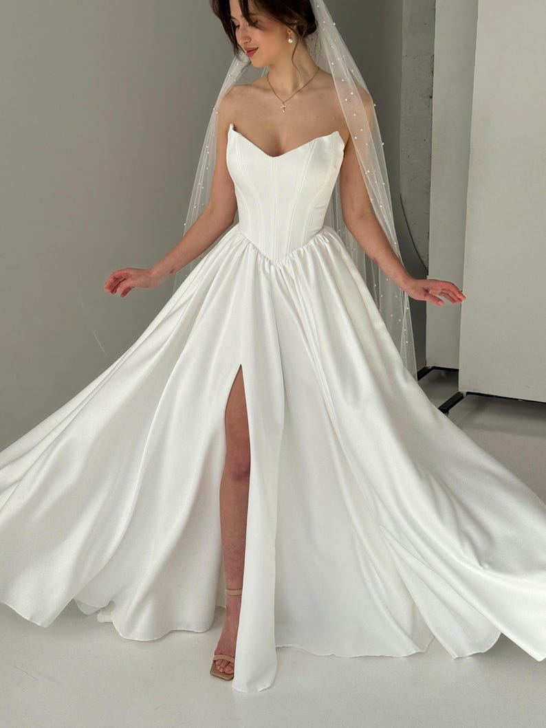 May include: A white strapless wedding dress with a sweetheart neckline and a thigh-high slit. The dress has a fitted bodice and a flowing skirt. The dress is paired with a white veil with pearl embellishments.