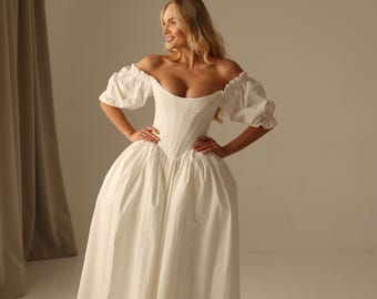 Könnte beinhalten: Ein bodenlanges weißes Kleid mit schulterfreiem Ausschnitt, kurzen Puffärmeln mit gerafften elastischen Bündchen und einem strukturierten Oberteil im Korsett-Stil, das in einen voluminösen, bodenlangen Rock mit glatter Oberfläche übergeht.