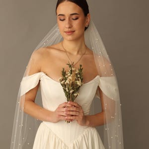 May include: A bride in an off-white, strapless wedding dress with a veil adorned with small pearls. She holds a small bouquet of dried flowers. The dress has a fitted bodice and a flowing skirt.