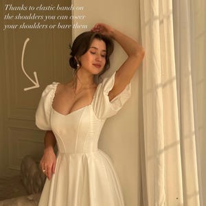 May include: Ivory satin a-line midi dress with puff sleeves and a sweetheart neckline.  The dress features elastic bands on the shoulders, allowing for adjustable shoulder coverage. The bodice is fitted with a corset-style seam detail.  The skirt flows into a full, gathered a-line shape.  The overall style is romantic and elegant.