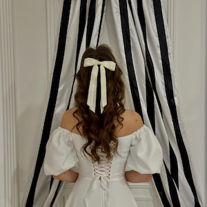 May include: A white off-the-shoulder dress with a lace-up back. The dress has puffy sleeves and a full skirt. The person is wearing a white bow in their hair.
