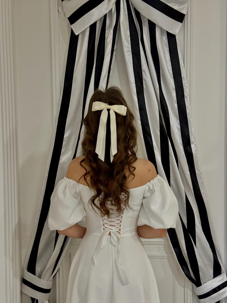 May include: A white off-the-shoulder dress with a lace-up back. The dress has puffy sleeves and a full skirt. The person is wearing a white bow in their hair.