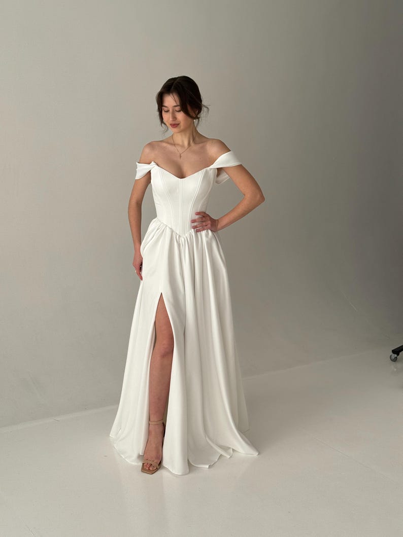 May include: A white off-the-shoulder wedding dress with a fitted bodice and a flowing skirt. The skirt has a high slit on the left side.