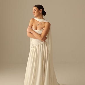 May include: A white wedding dress with a sweetheart neckline and a flowing skirt. The dress features a fitted bodice and a long train. A matching cape drapes over the shoulders. A silver necklace with a pendant is visible.