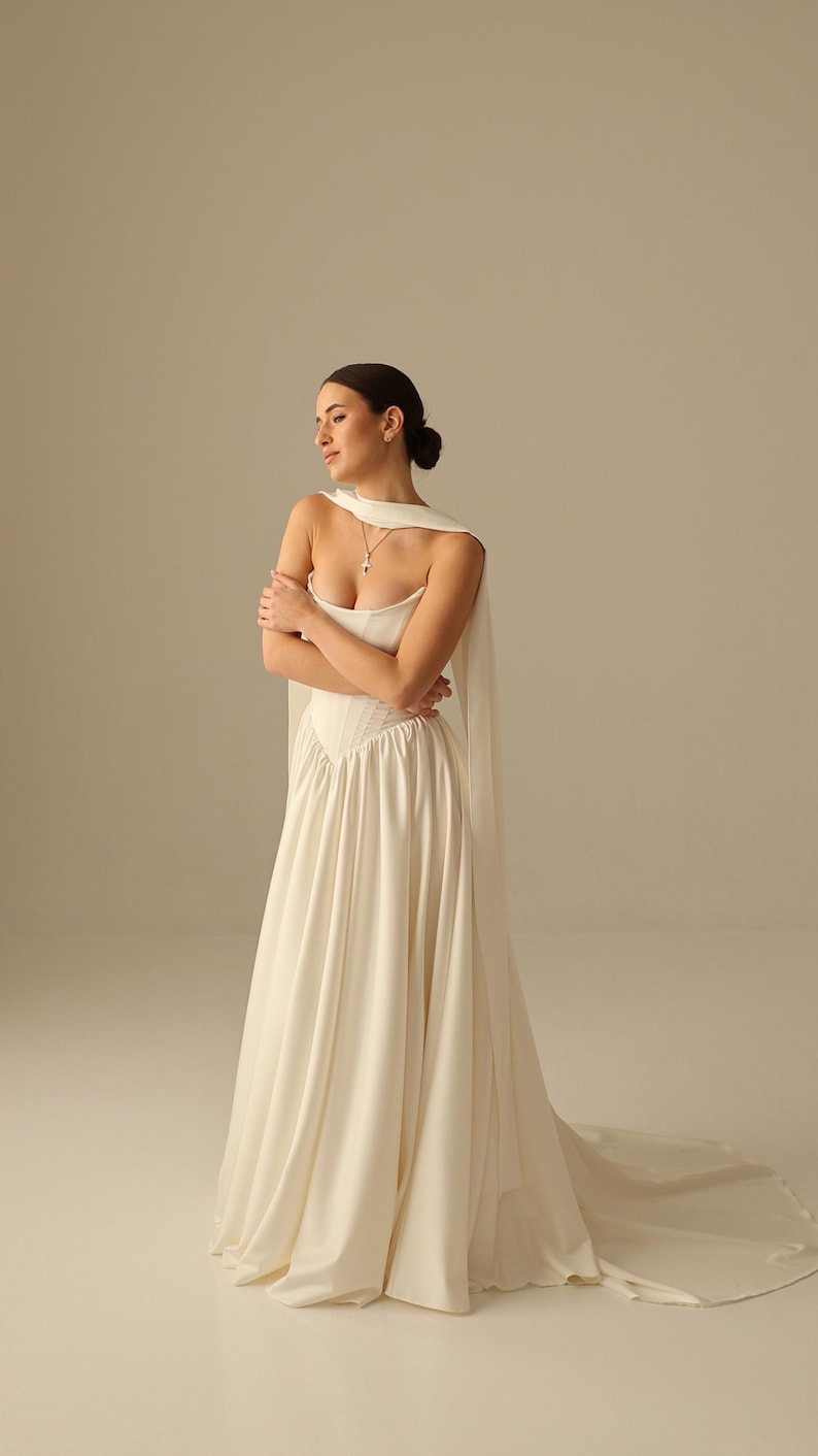 May include: A white wedding dress with a sweetheart neckline and a flowing skirt. The dress features a fitted bodice and a long train. A matching cape drapes over the shoulders. A silver necklace with a pendant is visible.