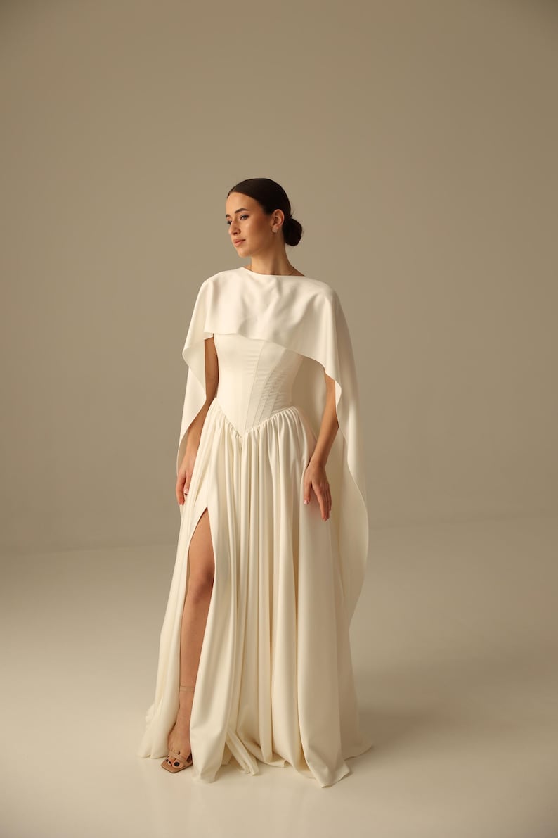 May include: An ivory-colored bridal gown with a high slit and a matching cape. The dress features a fitted bodice and a flowing skirt. The model is wearing beige sandals. The overall style is elegant and sophisticated, suitable for a wedding or formal event.
