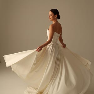 May include: A strapless ivory wedding dress with a fitted bodice and a flowing skirt. The dress features a lace-up back and is made of a smooth, satin-like fabric. The bride is twirling, showing the dress's movement.