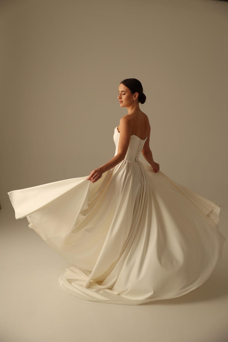 May include: A strapless ivory wedding dress with a fitted bodice and a flowing skirt. The dress features a lace-up back and is made of a smooth, satin-like fabric. The bride is twirling, showing the dress's movement.