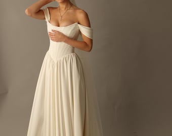 May include: An off-white wedding dress with an off-the-shoulder neckline and a fitted bodice. The dress has a full skirt that flows to the floor, and a long train. A veil with small pearls is attached to the back of the dress. The model is smiling.