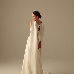 May include: A white bridal gown with a flowing skirt and a cape-like overlay. The dress features a keyhole back and a long train. The model is turned to the side, showcasing the dress's design and silhouette. The overall aesthetic is elegant and minimalist.