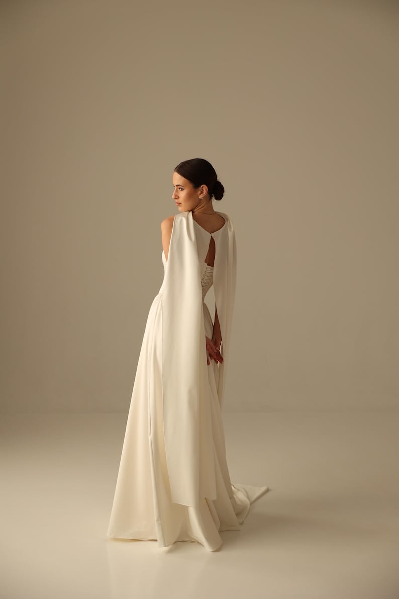 May include: A white bridal gown with a flowing skirt and a cape-like overlay. The dress features a keyhole back and a long train. The model is turned to the side, showcasing the dress's design and silhouette. The overall aesthetic is elegant and minimalist.