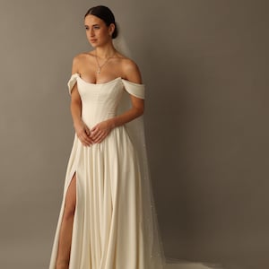 May include: An off-white wedding dress with an off-the-shoulder neckline and a high slit. The dress has a fitted bodice and a flowing skirt. A sheer veil with pearl accents trails behind the dress. The model is wearing a necklace and sandals.