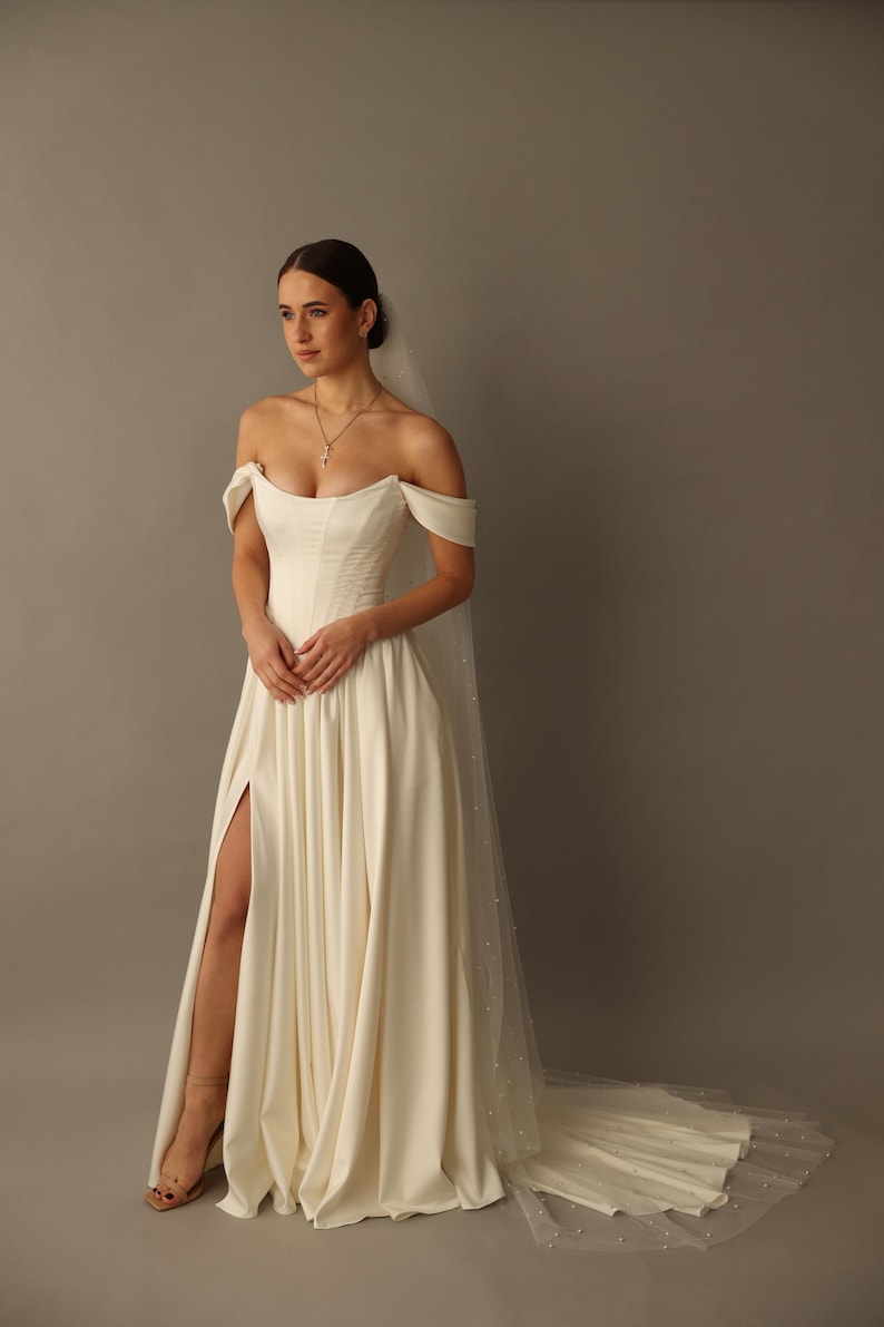 May include: An off-white wedding dress with an off-the-shoulder neckline and a high slit. The dress has a fitted bodice and a flowing skirt. A sheer veil with pearl accents trails behind the dress. The model is wearing a necklace and sandals.
