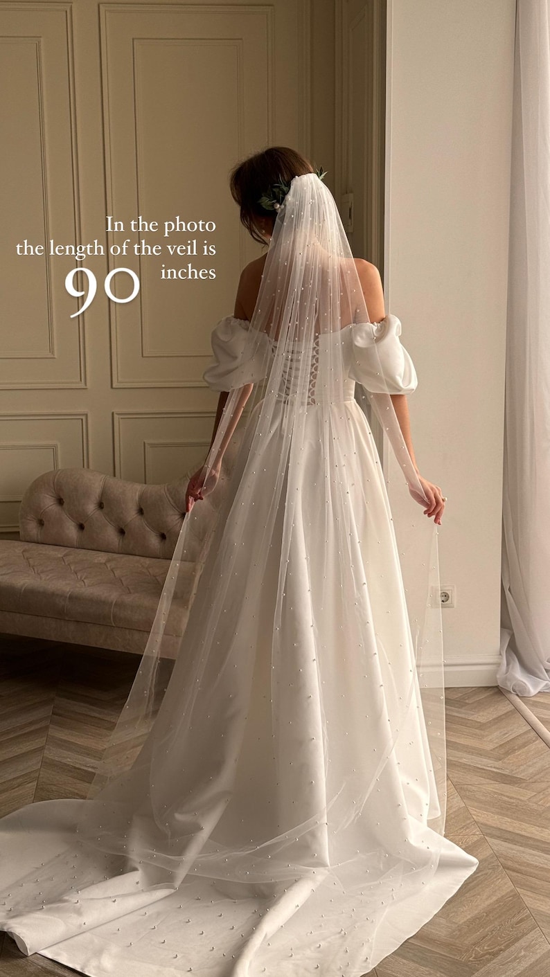 May include: A floor-length wedding veil, 90 inches long, with scattered pearl embellishments. The veil is made of ivory tulle and is shown draped over a satin wedding gown with puff sleeves and a lace-up back.  The veil is sheer and delicate, with the pearls adding subtle sparkle. The overall style is elegant and romantic.