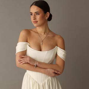 Pu&ograve; includere: Abito da sposa avorio con spalle scoperte, corpetto aderente e gonna fluida. L'abito presenta uno scollo a cuore ed &egrave; impreziosito da una collana con pendente a croce argentata e un braccialetto. Le braccia della modella sono incrociate.
