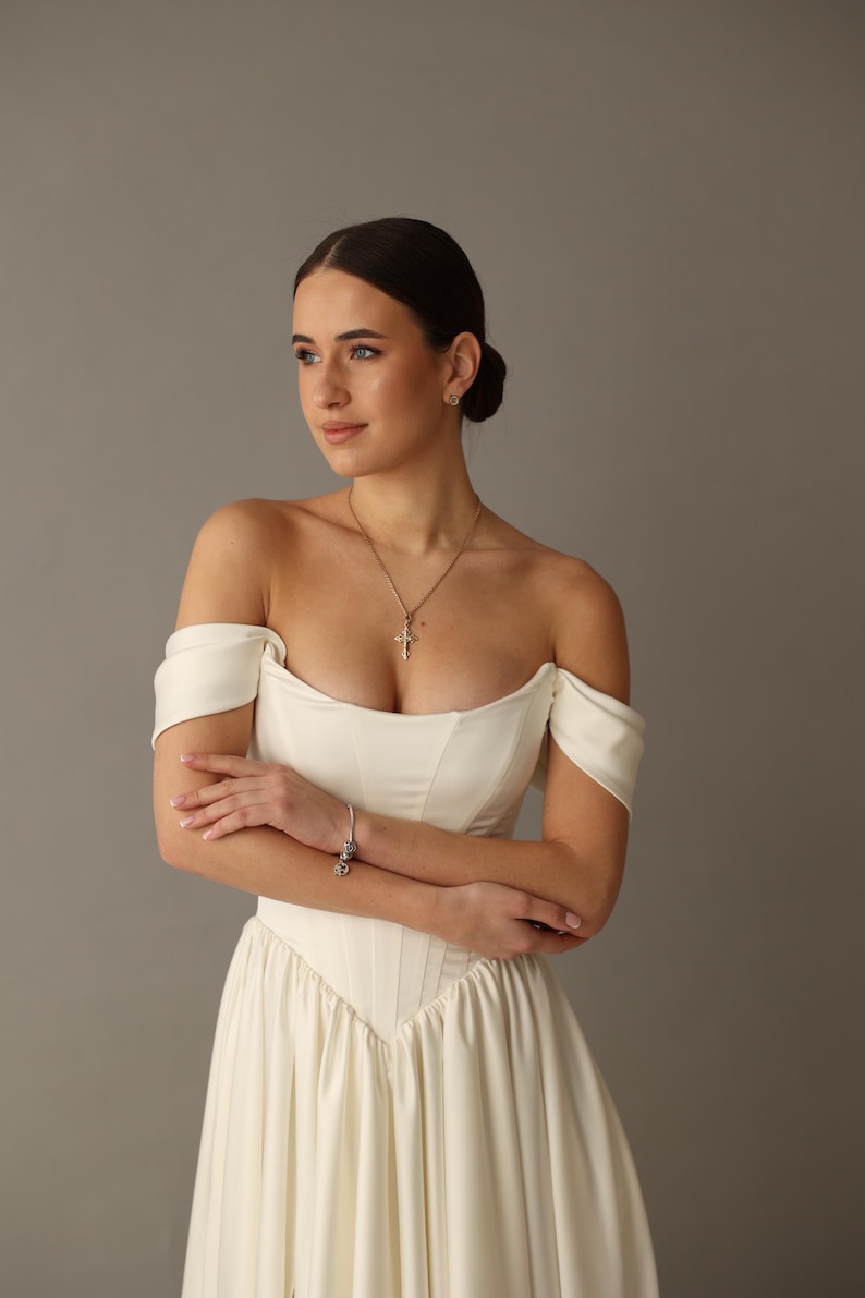 Pu&ograve; includere: Abito da sposa avorio con spalle scoperte, corpetto aderente e gonna fluida. L'abito presenta uno scollo a cuore ed &egrave; impreziosito da una collana con pendente a croce argentata e un braccialetto. Le braccia della modella sono incrociate.