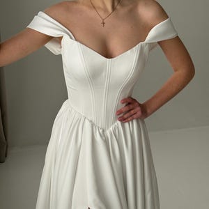 May include: A white off-the-shoulder wedding dress with a sweetheart neckline and a fitted bodice. The skirt is flowing and has a thigh-high slit.