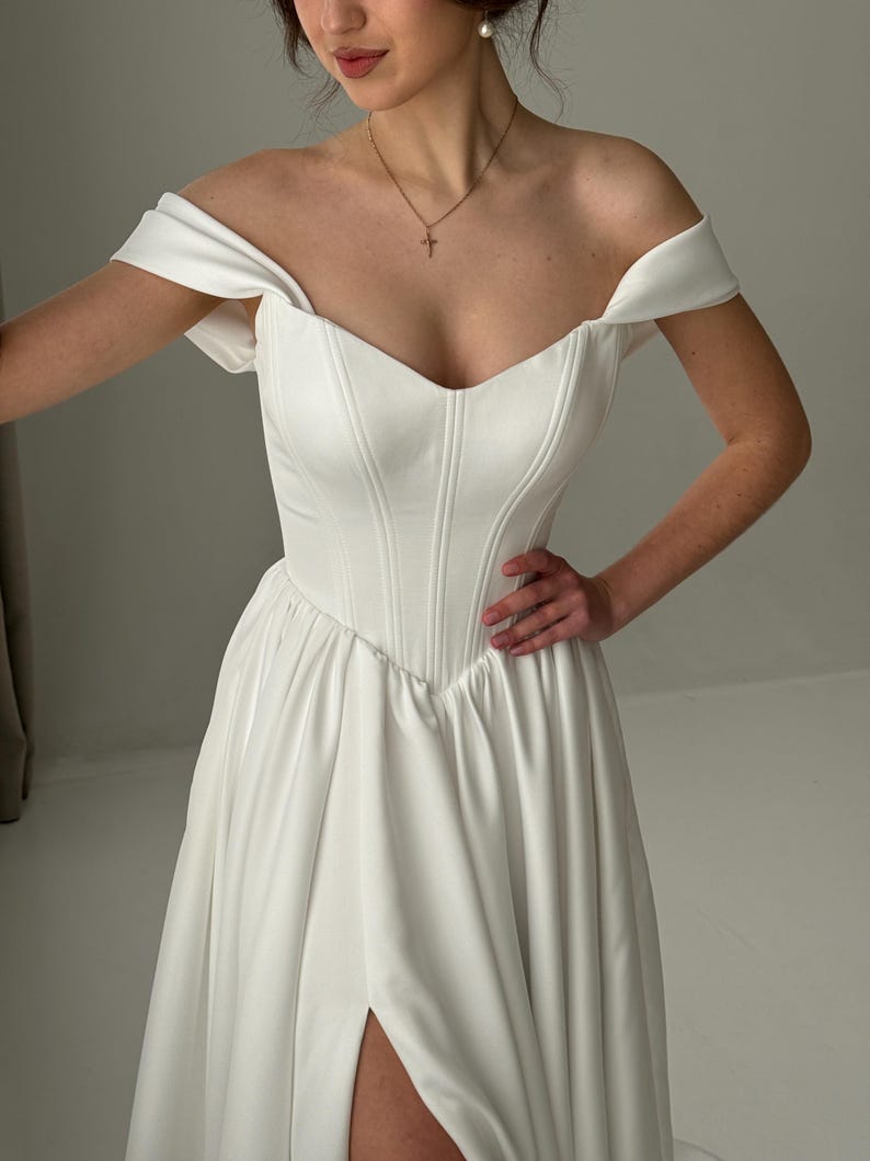 May include: A white off-the-shoulder wedding dress with a sweetheart neckline and a fitted bodice. The skirt is flowing and has a thigh-high slit.