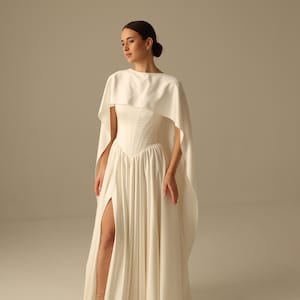Pu&ograve; includere: Abito da sposa color avorio con mantella, con corpetto aderente e gonna fluida con spacco alto. L'abito &egrave; abbinato a sandali. Lo stile generale &egrave; elegante e sofisticato, adatto per un matrimonio o un'occasione speciale.