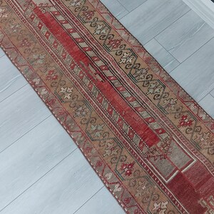 May include: A rectangular, vintage runner rug with a central red stripe and intricate geometric patterns in beige, brown, and gray. The rug features a distressed look, with faded colors and a worn texture, suggesting age and use. The rug is suitable for a hallway or entryway.